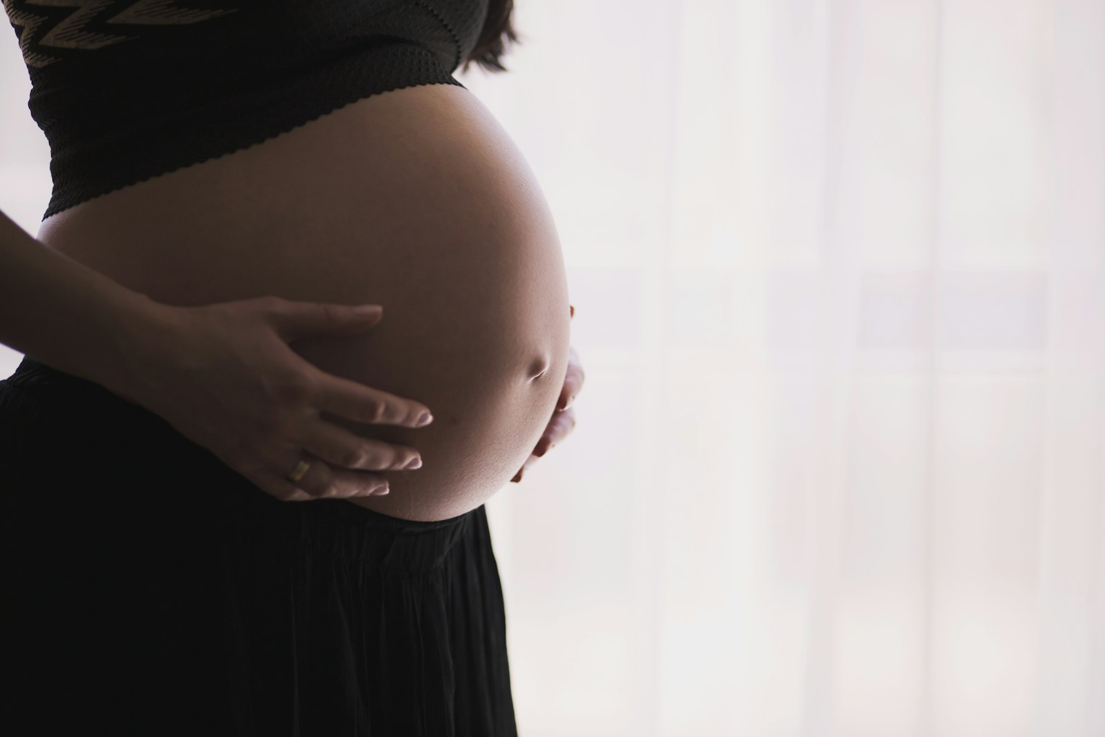 A side view of a pregnant belly with a hand resting on it, soft window light behind.