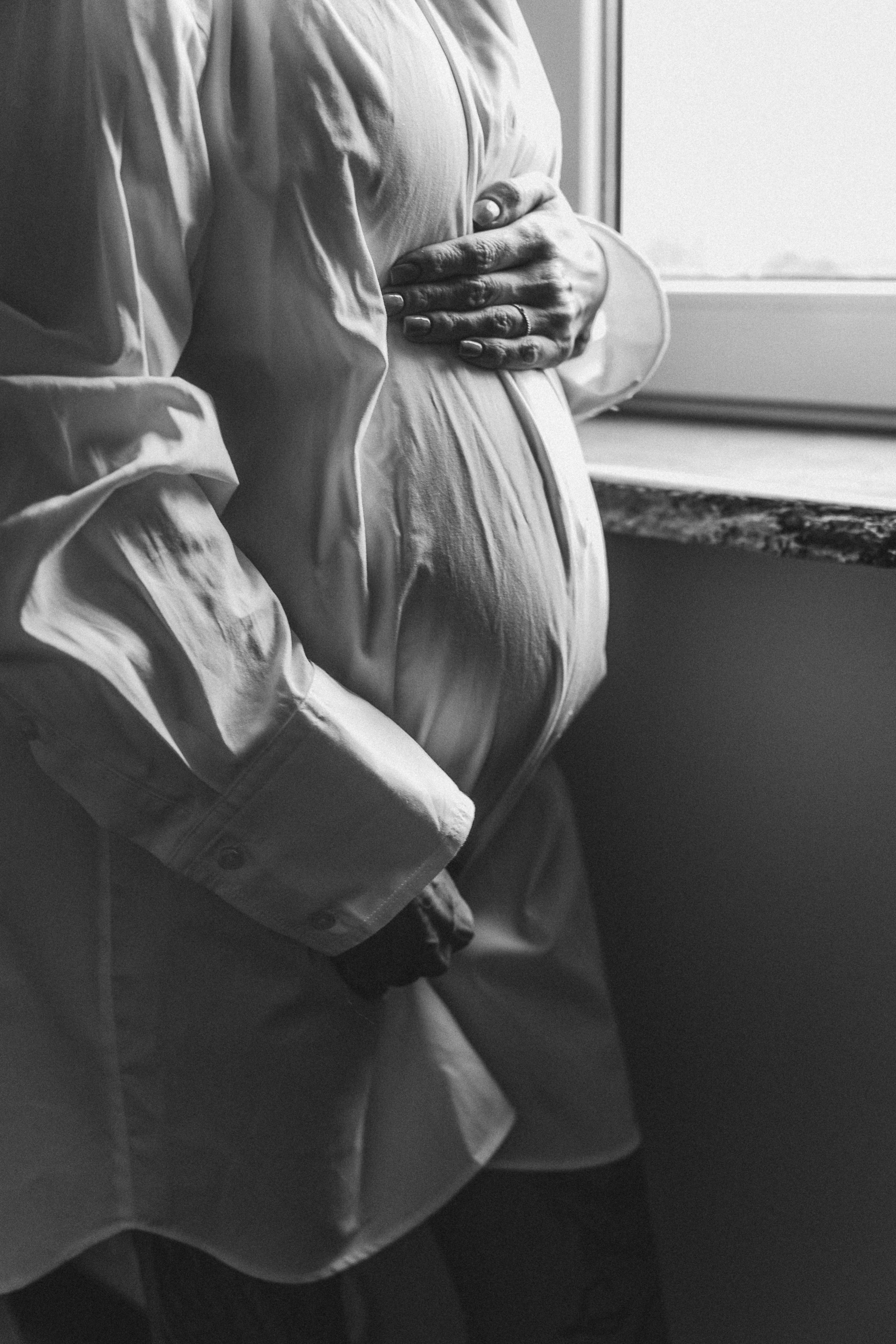 Black-and-white photo of a pregnant woman in a soft linen shirt, one hand resting gently on her belly, standing by a window.